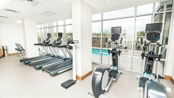 A gym with treadmills, elliptical machines, and a rowing machine, large windows, and a view of a pool.