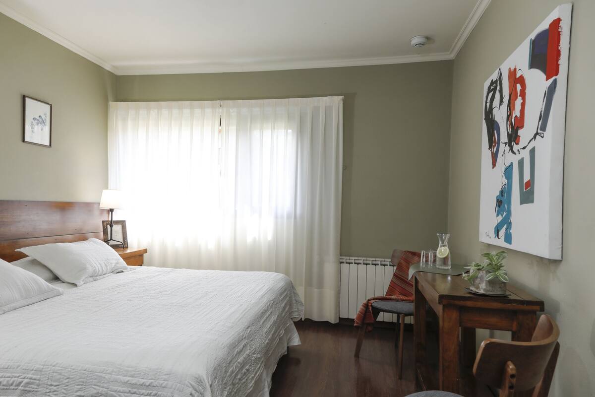 Quarto de hotel com uma cama grande, uma escrivaninha com cadeira, um radiador e um quadro na parede.
