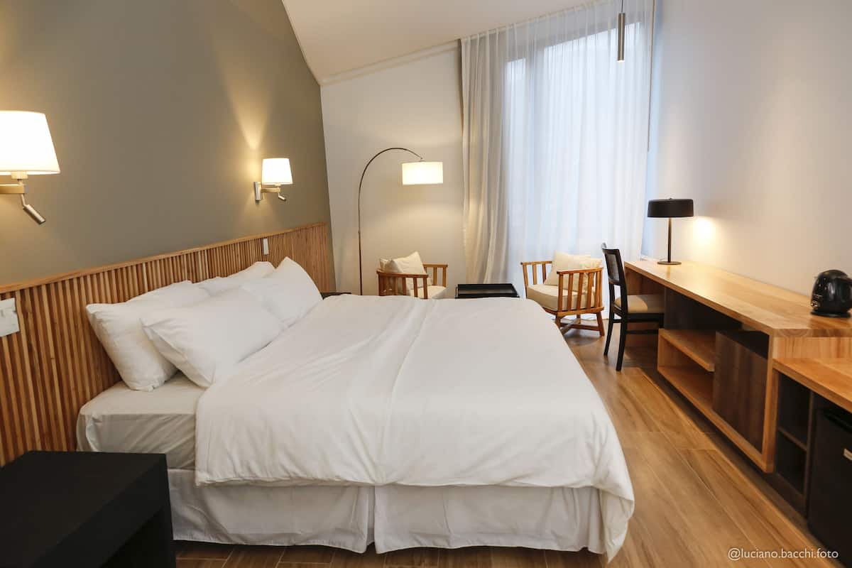 Quarto de hotel com uma cama grande, uma mesa com cadeira, uma luminária e vista para o exterior.
