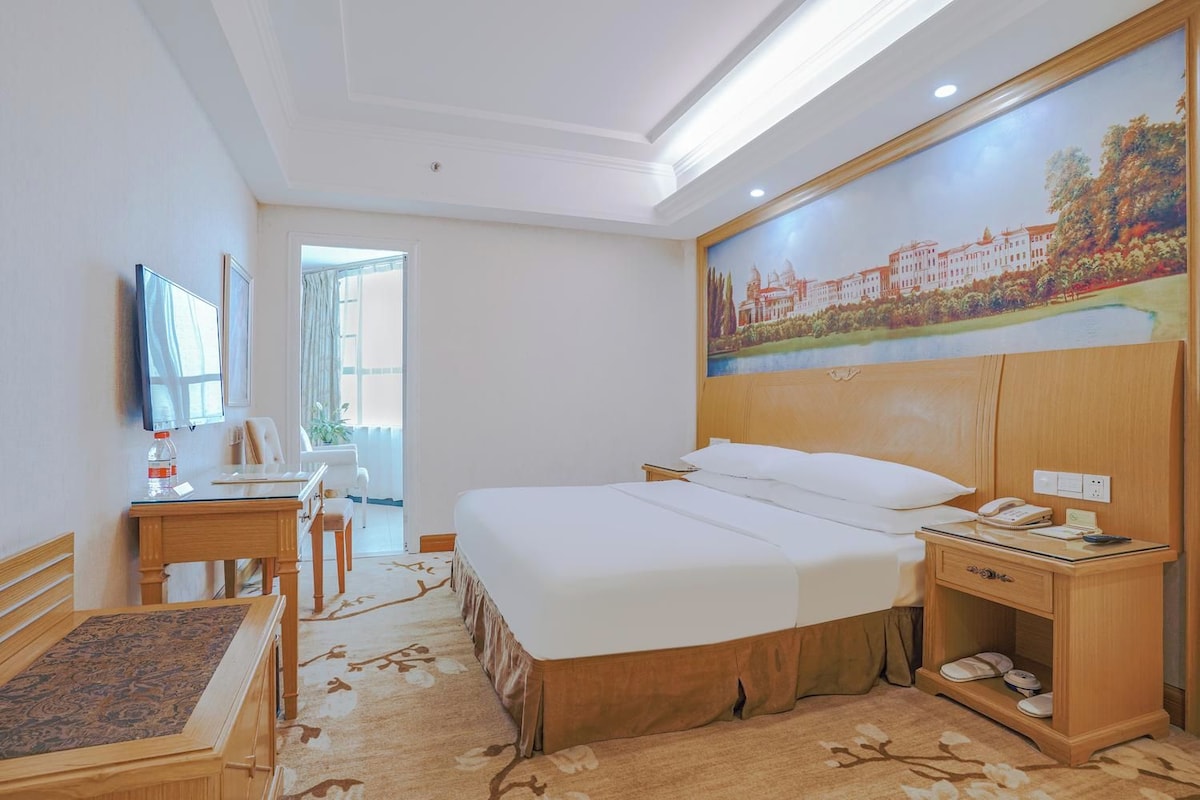 A hotel room with a large bed, a desk with a chair, a television, a painting on the wall, and a window with curtains.