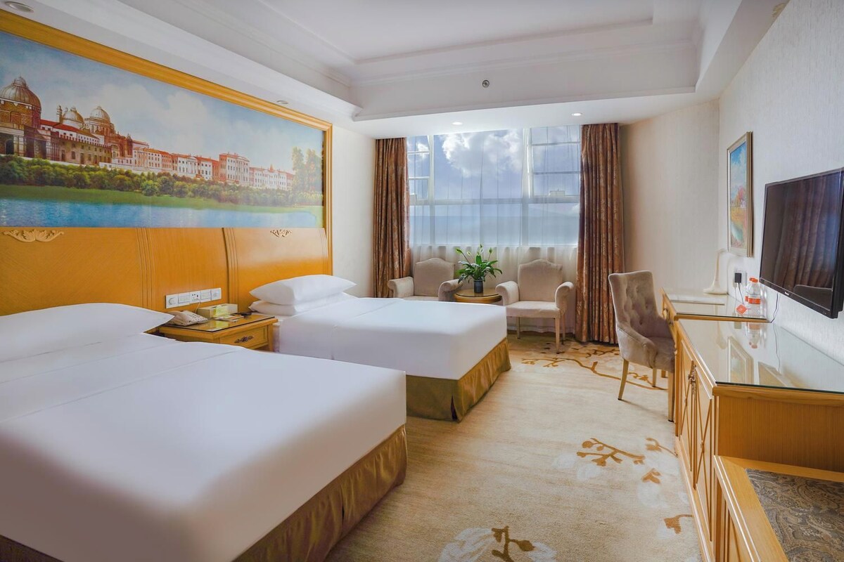 A hotel room with two beds, a desk with a TV, a chair, a window with curtains, and a painting on the wall.