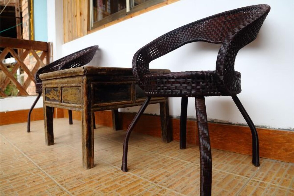 Two wicker chairs with wooden legs and a wooden table, placed on a tiled floor against a white wall.