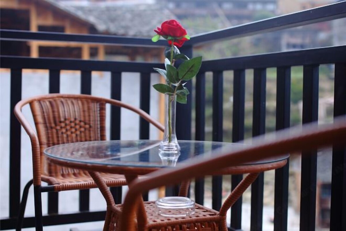A balcony with wicker chairs and a glass table, a vase with a single red rose, and a view of buildings in the background.