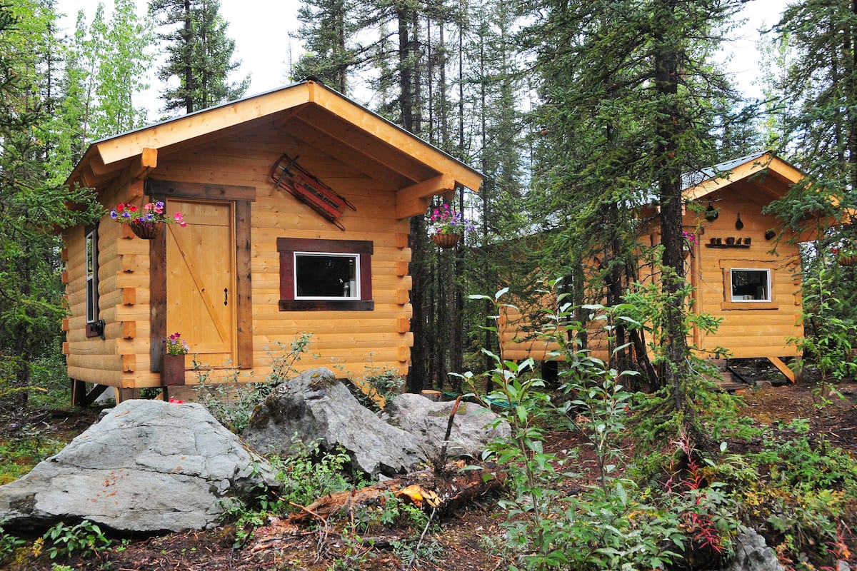 Deux cabanes en bois, entourées de forêt. Chaque cabane est dotée d’une porte, de fenêtres et d’un petit porche.