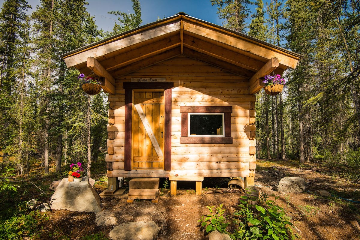 Une petite cabane en bois, dotée d’un toit en pente, d’une fenêtre et d’une porte. Des jardinières sont suspendues aux murs extérieurs.