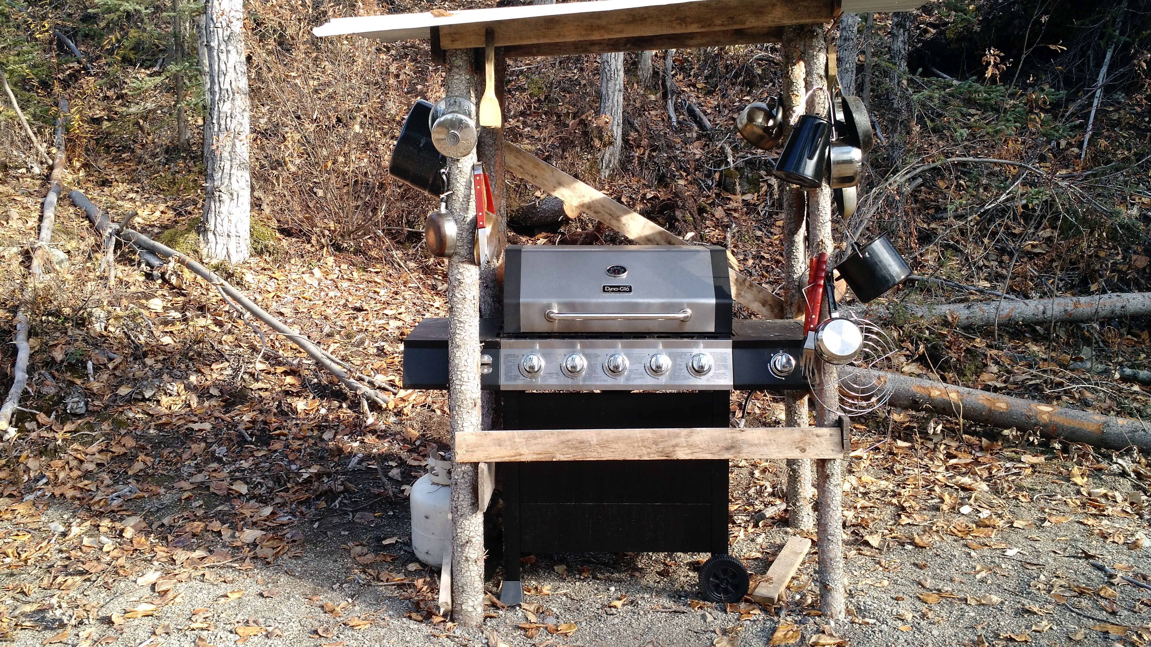 Una parrilla portátil con cuatro quemadores, un quemador lateral y un compartimento de almacenamiento, montada al aire libre sobre una estructura de madera y rodeada de árboles y hojas secas.