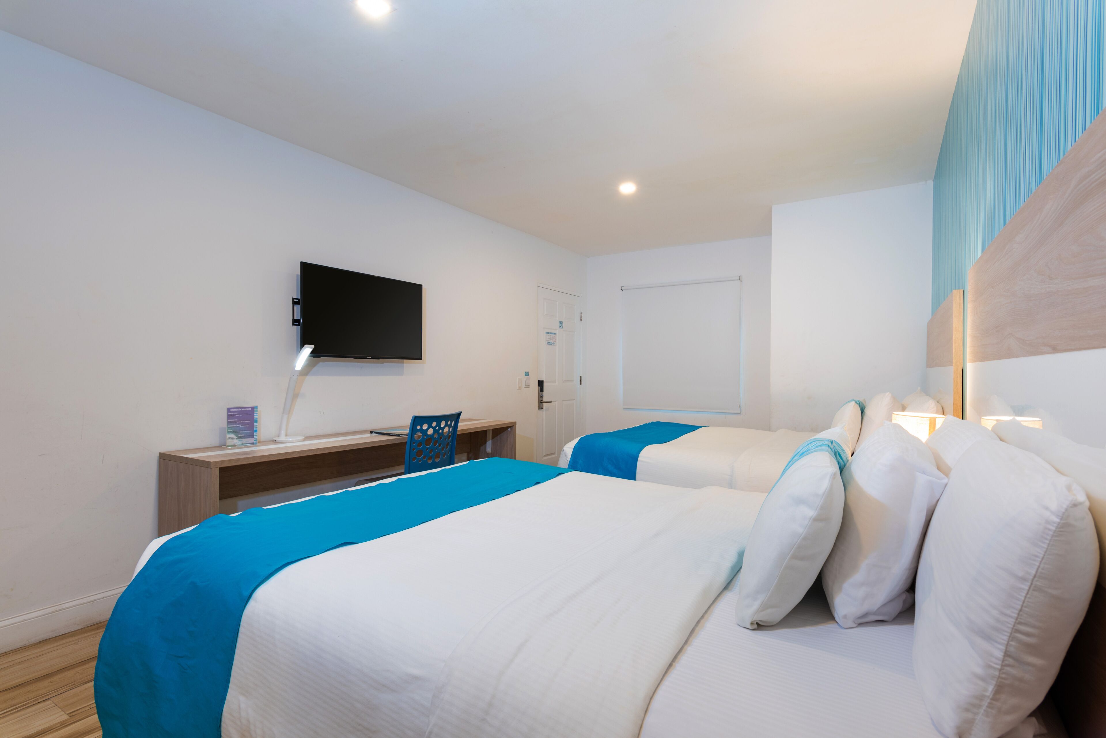 A hotel room with two beds, a desk, a chair, a TV, and a door.