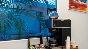 Una máquina de café con el logotipo de la marca Bunn, una bandeja con vasos y cubiertos desechables, y una caja de aperitivos sobre una encimera cerca de una ventana con vistas de color azulado.