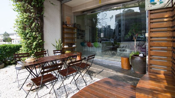 Un espacio al aire libre con mesa y sillas de madera, rodeado de follaje y un muro de cristal.