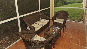 Un patio couvert aménagé avec des meubles en osier, notamment un canapé et deux fauteuils, ainsi qu’une table basse en verre.