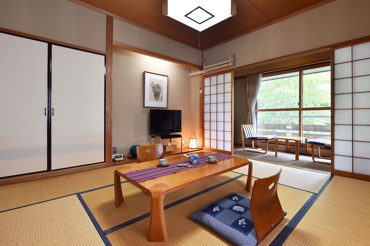 Una habitación tradicional japonesa con suelo de tatami, una mesa de madera baja, un televisor y puertas correderas que dan a un balcón con vistas a la vegetación.