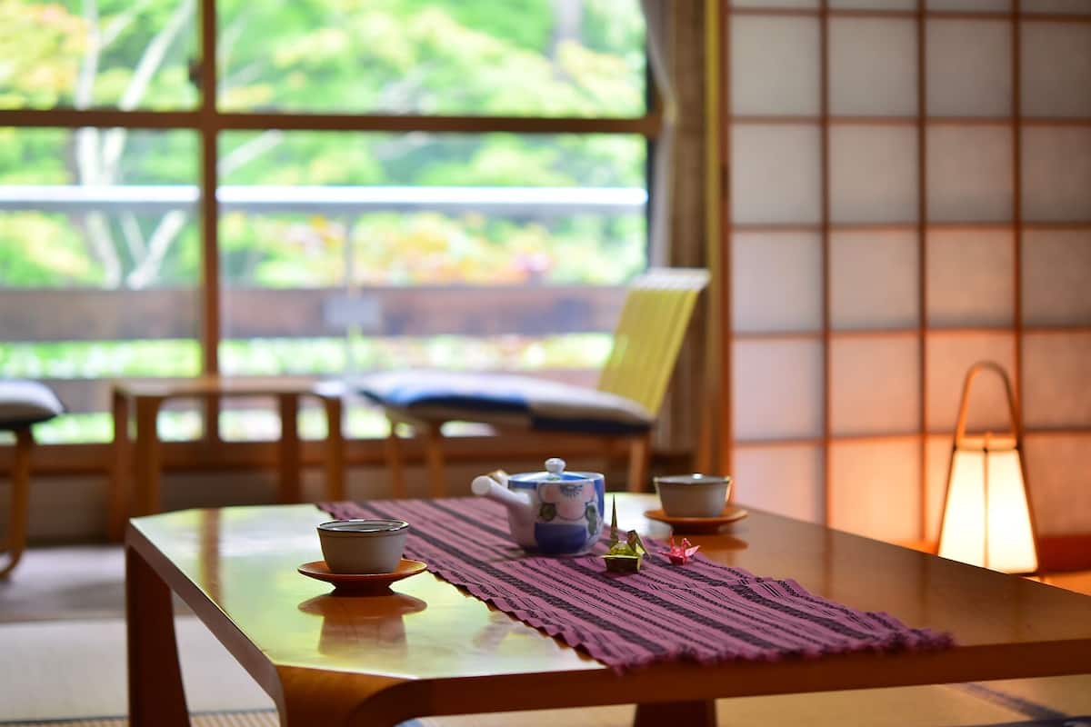Una habitación japonesa tradicional con suelo de tatami, una mesa baja con un juego de té, una silla y vistas a través de un celosón.
