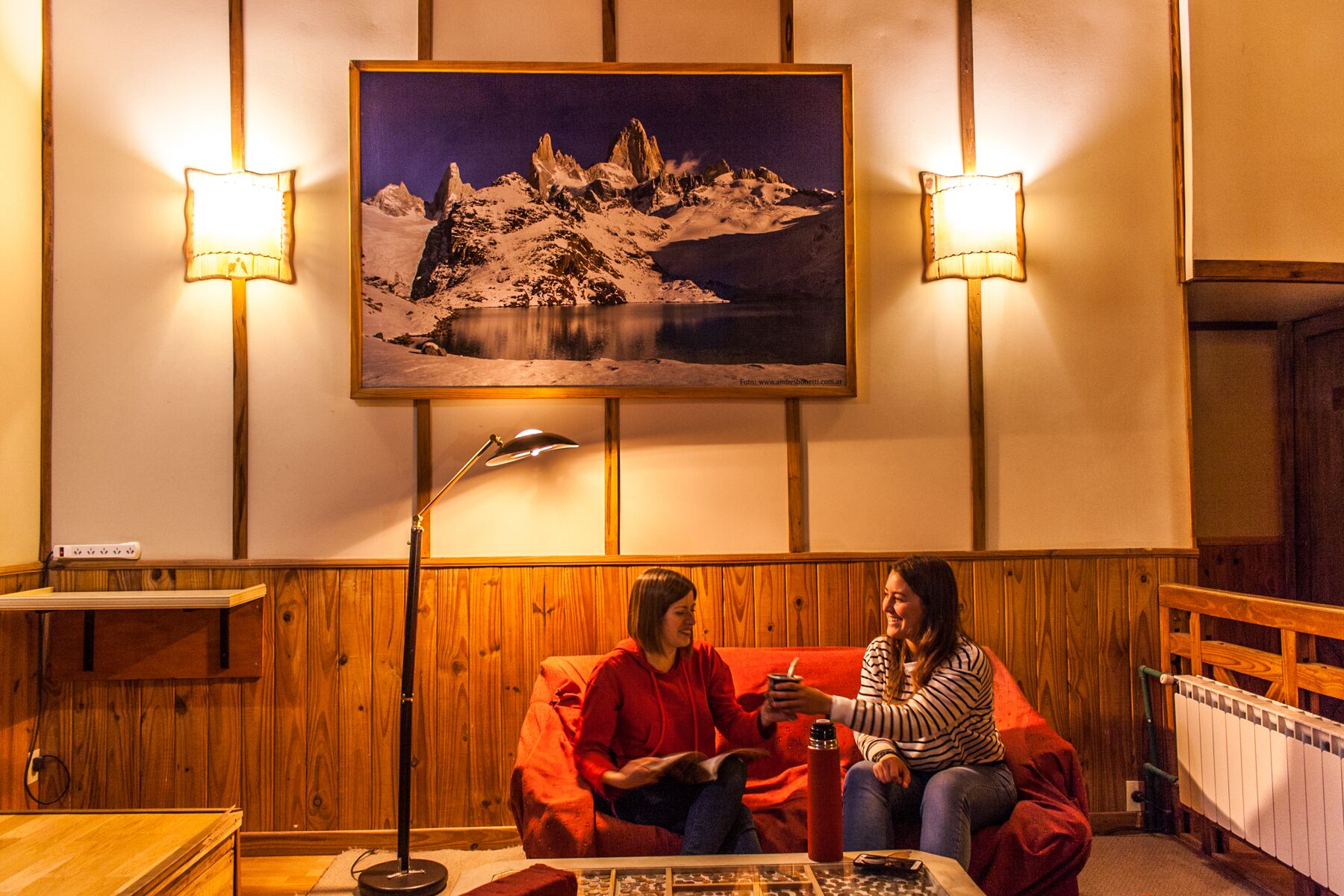 A cozy room with wooden paneling, a large painting of a mountain landscape, and two people seated on a couch.