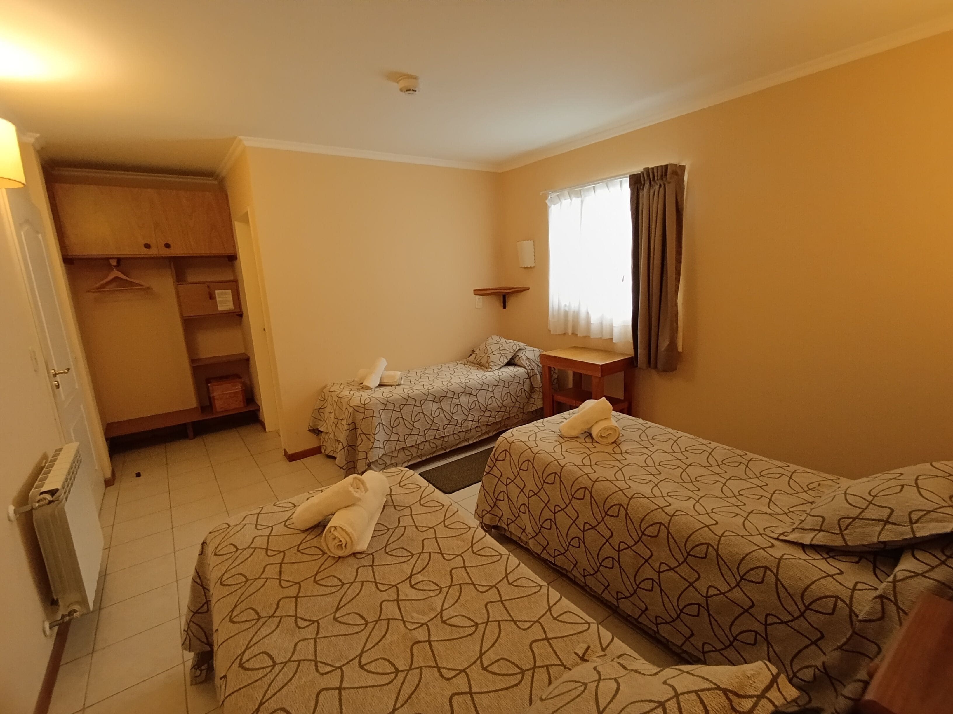 Una habitación de hotel con dos camas, un armario, una mesita y una ventana con cortinas.
