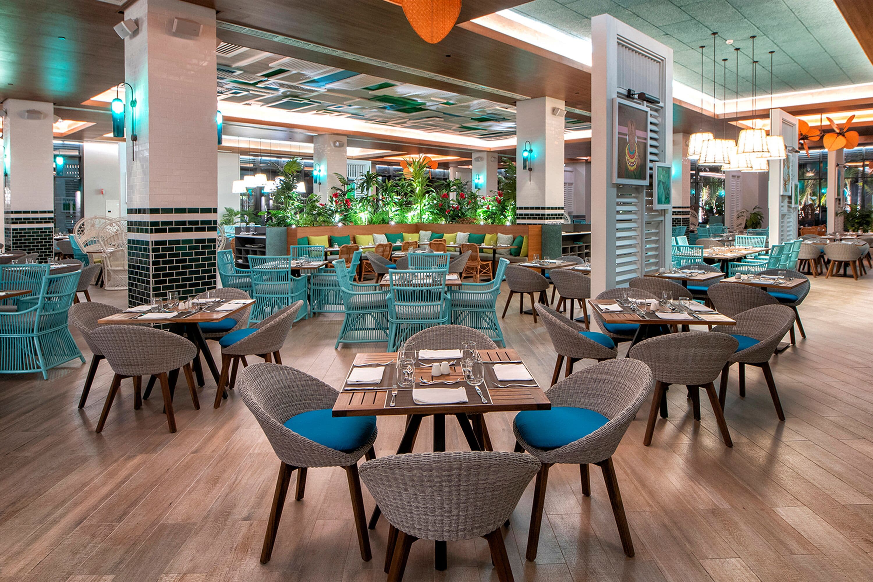 Ein Restaurant mit Korbstühlen, blauen Kissen, Holztischen und einer zentralen Sitzgruppe mit einer niedrigen Mauer und Pflanzen.