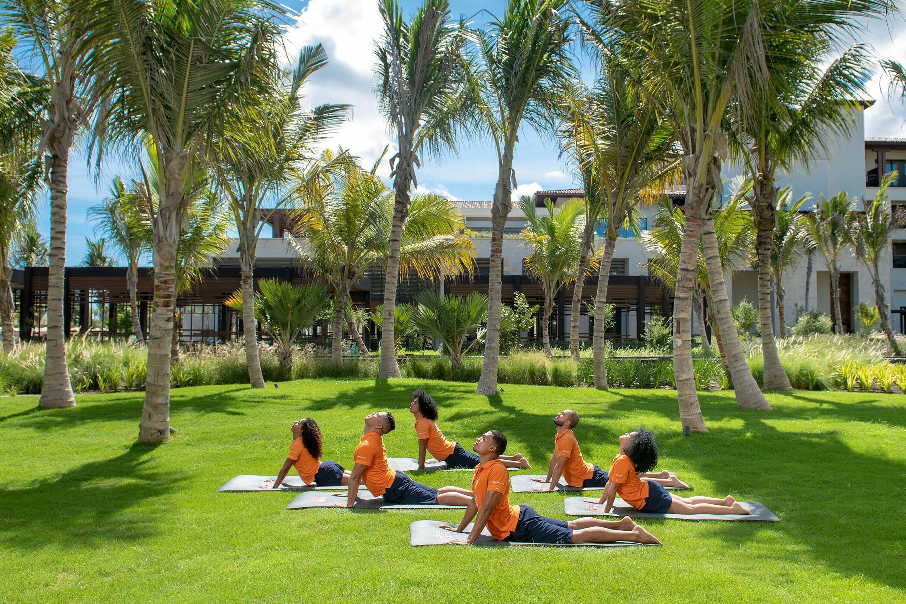 Un gruppo di persone che pratica yoga su tappetini in una zona erbosa con palme e un edificio sullo sfondo.