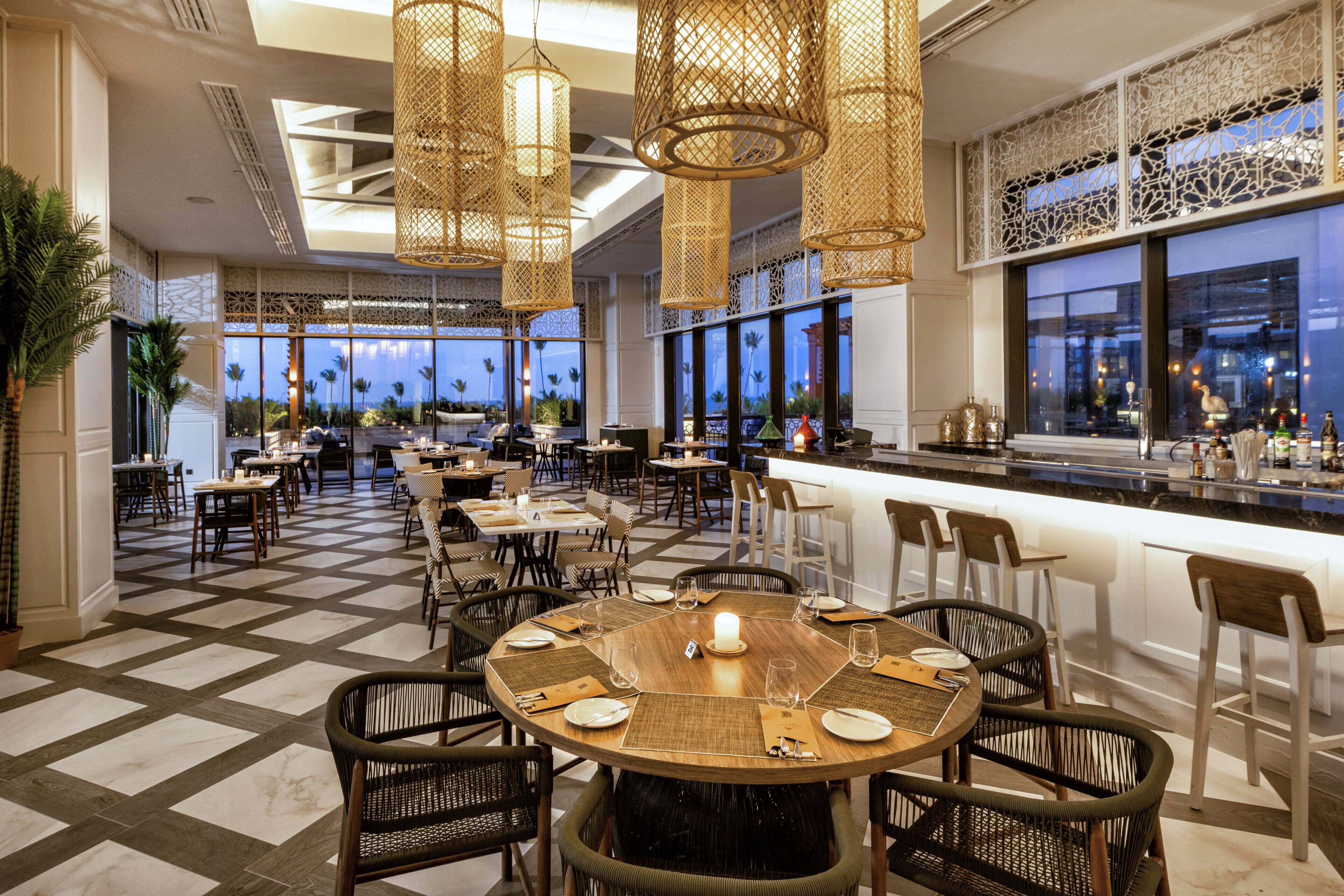 A restaurant with round tables, wicker chairs, and hanging light fixtures.