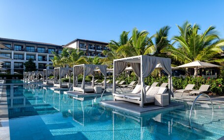 A pool area with lounge chairs and umbrellas, surrounded by palm trees and a building in the background.