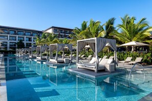 Un área de piscina con sillones de descanso y sombrillas, rodeada de palmeras y un edificio al fondo.