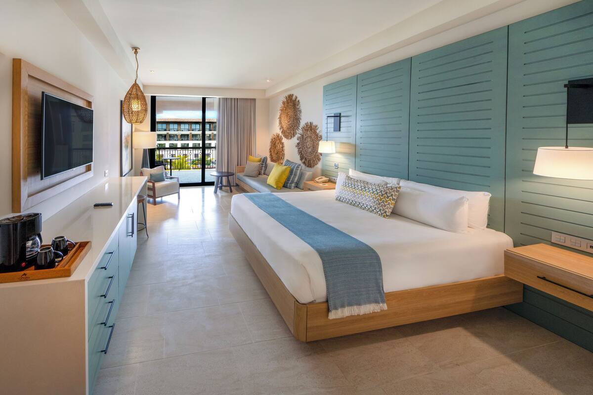 A modern hotel room with a large bed, a TV, a dining area, and a view of the outside.