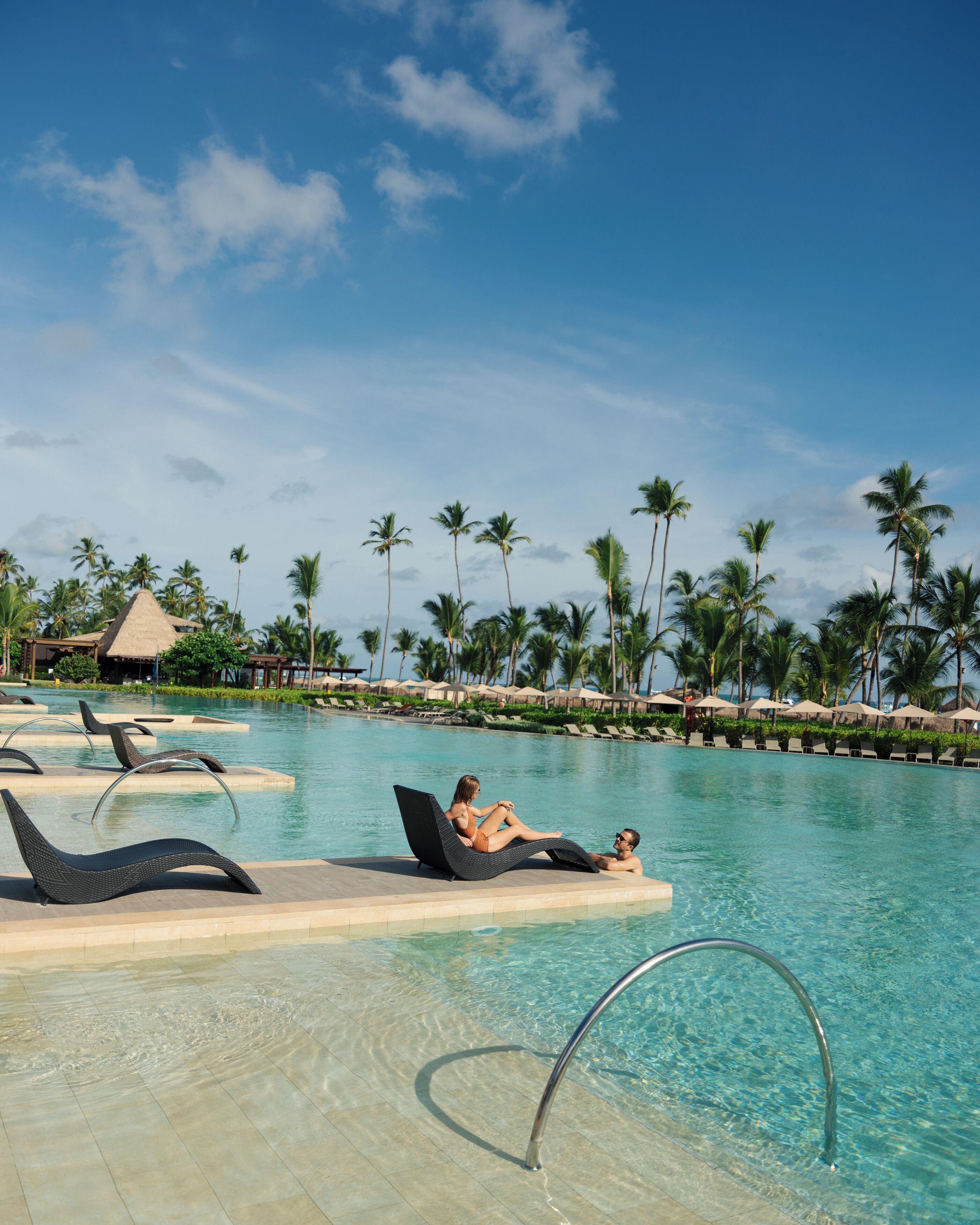 A poolside area with lounge chairs, a clear pool, palm trees, and a thatched-roof structure.