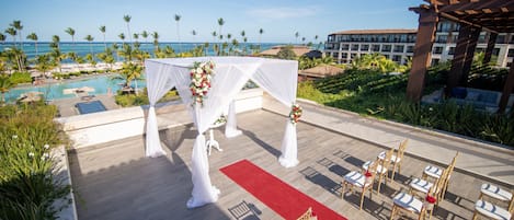 An outdoor wedding ceremony setup with a red carpet, white drapes, and floral arrangements.
