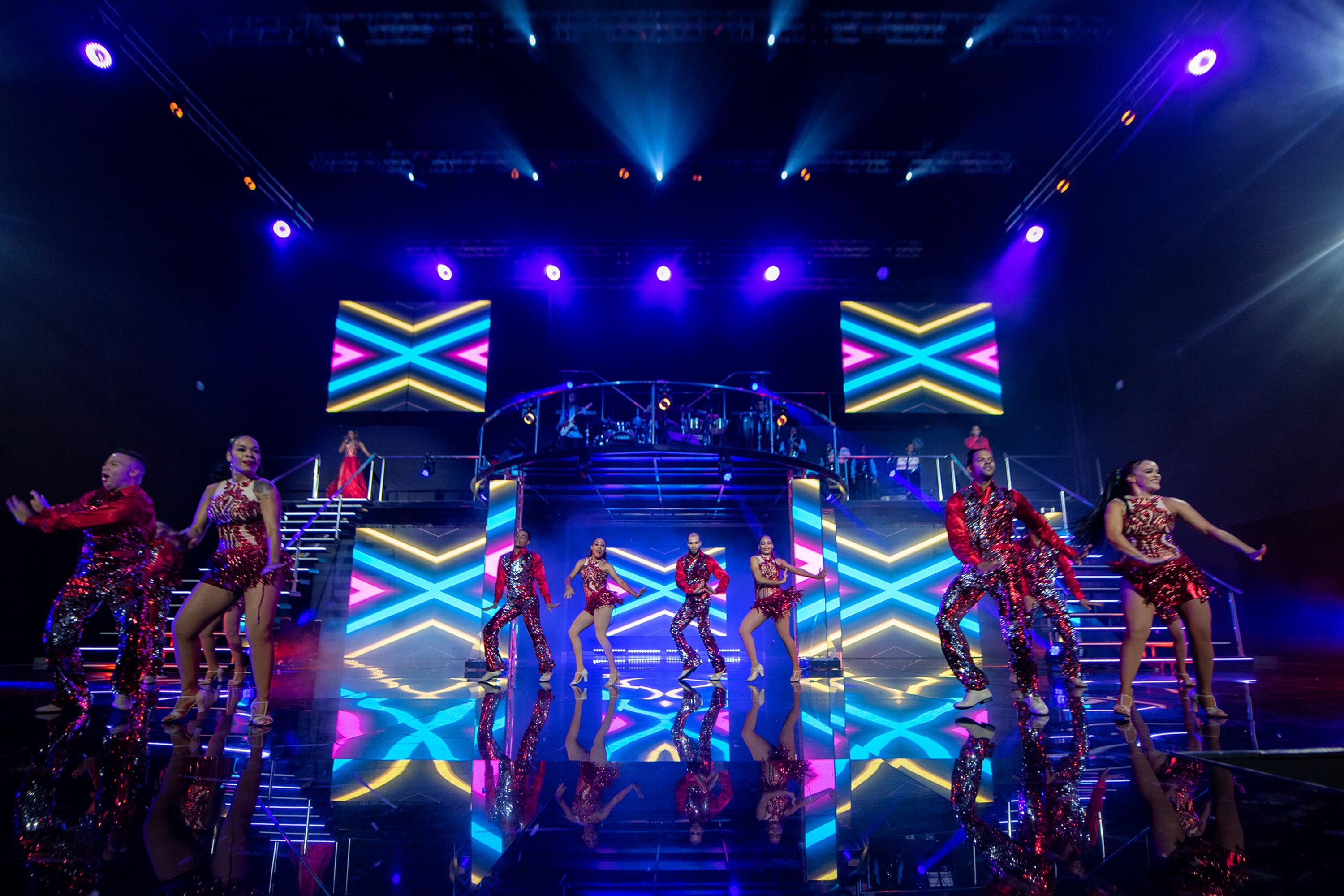 Une scène où des artistes portent des costumes rouges et argentés, un grand écran LED affichant un motif géométrique et un éclairage de scène vif.