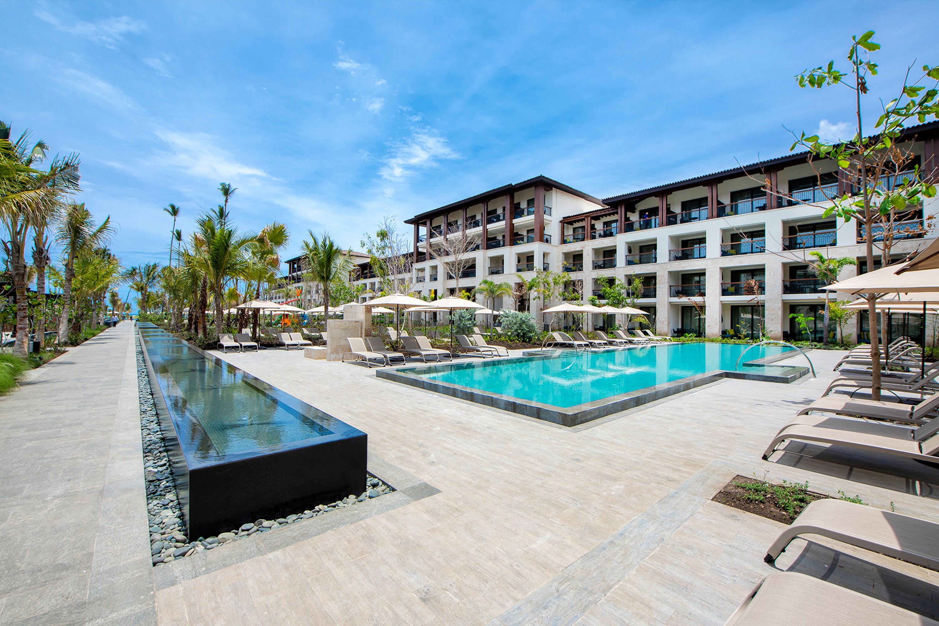 A resort pool area with a long rectangular pool, lounge chairs, and a building with multiple balconies.