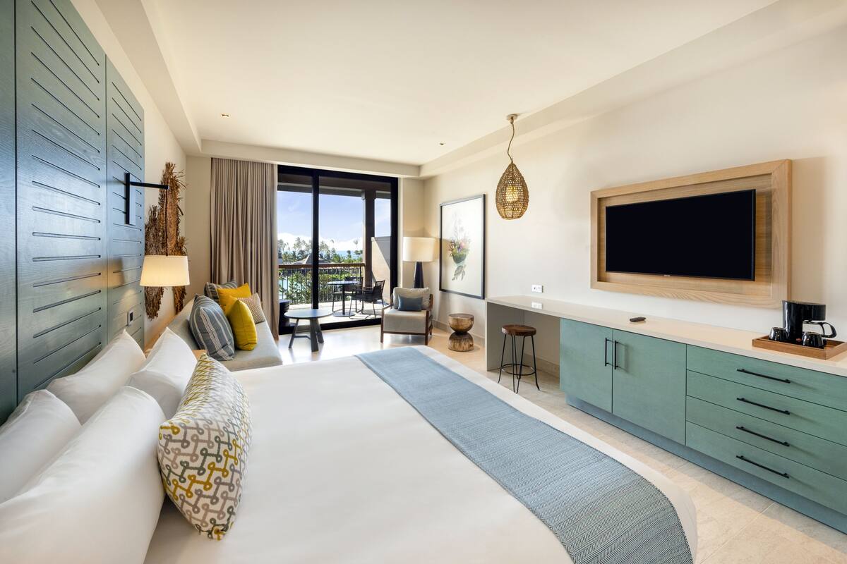A modern hotel room with a large bed, a balcony with a view, a kitchen area, and a flat-screen TV.