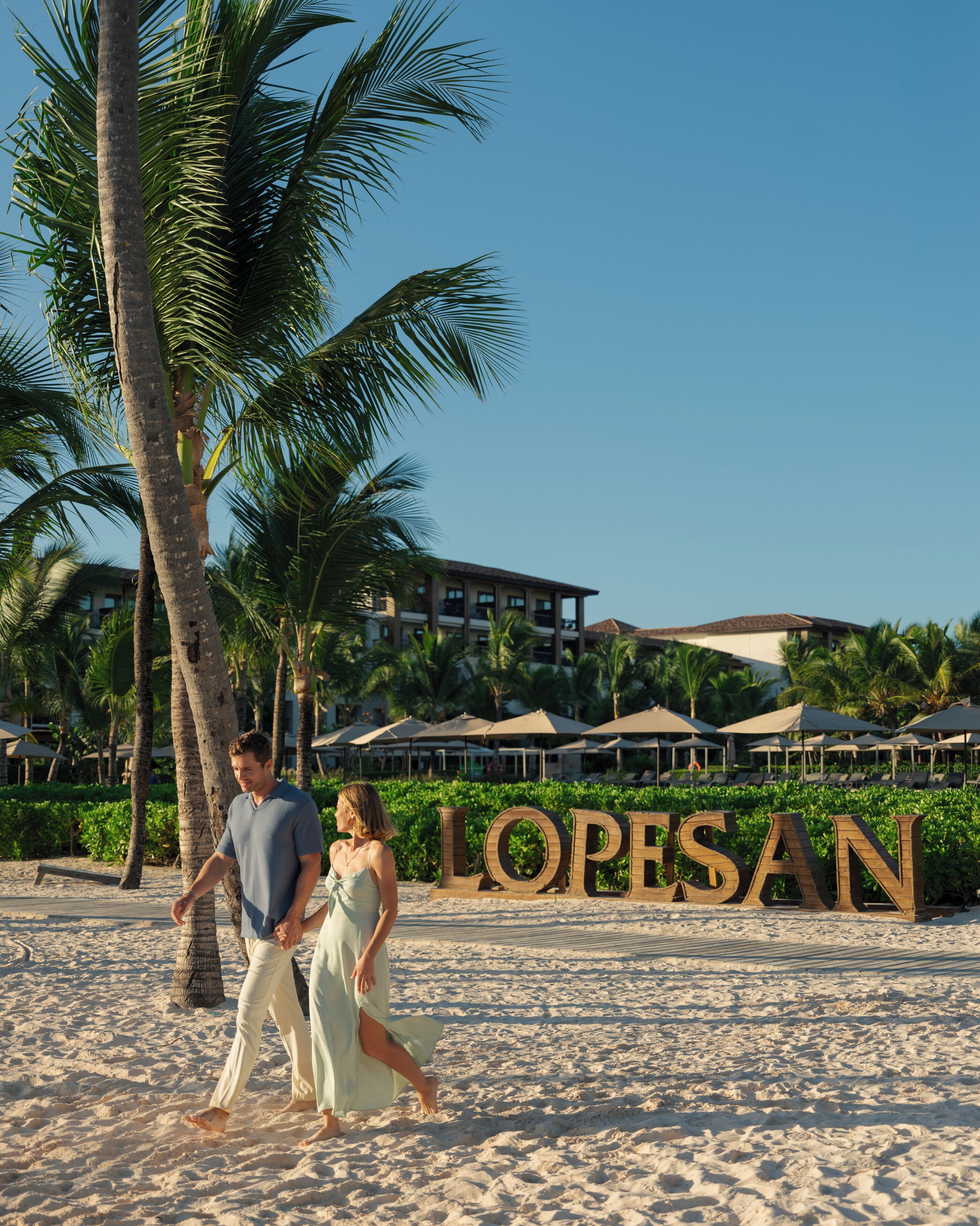A beach with palm trees and a resort named "LOPESAN" in the background.