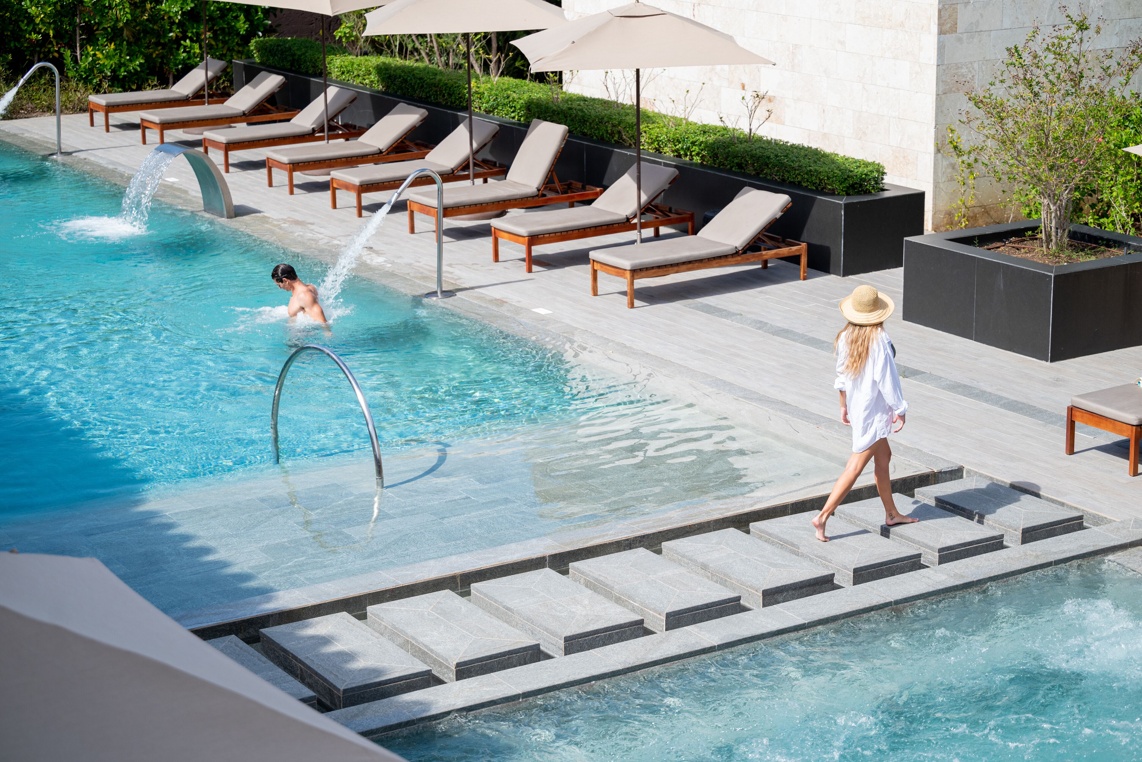 Zona de piscina con sillones reclinables, fuente de agua y planta en maceta.
