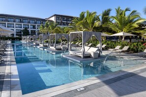 Ein Swimmingpool mit Liegestühlen und Sonnenschirmen, umgeben von Palmen und einem Hotelgebäude.