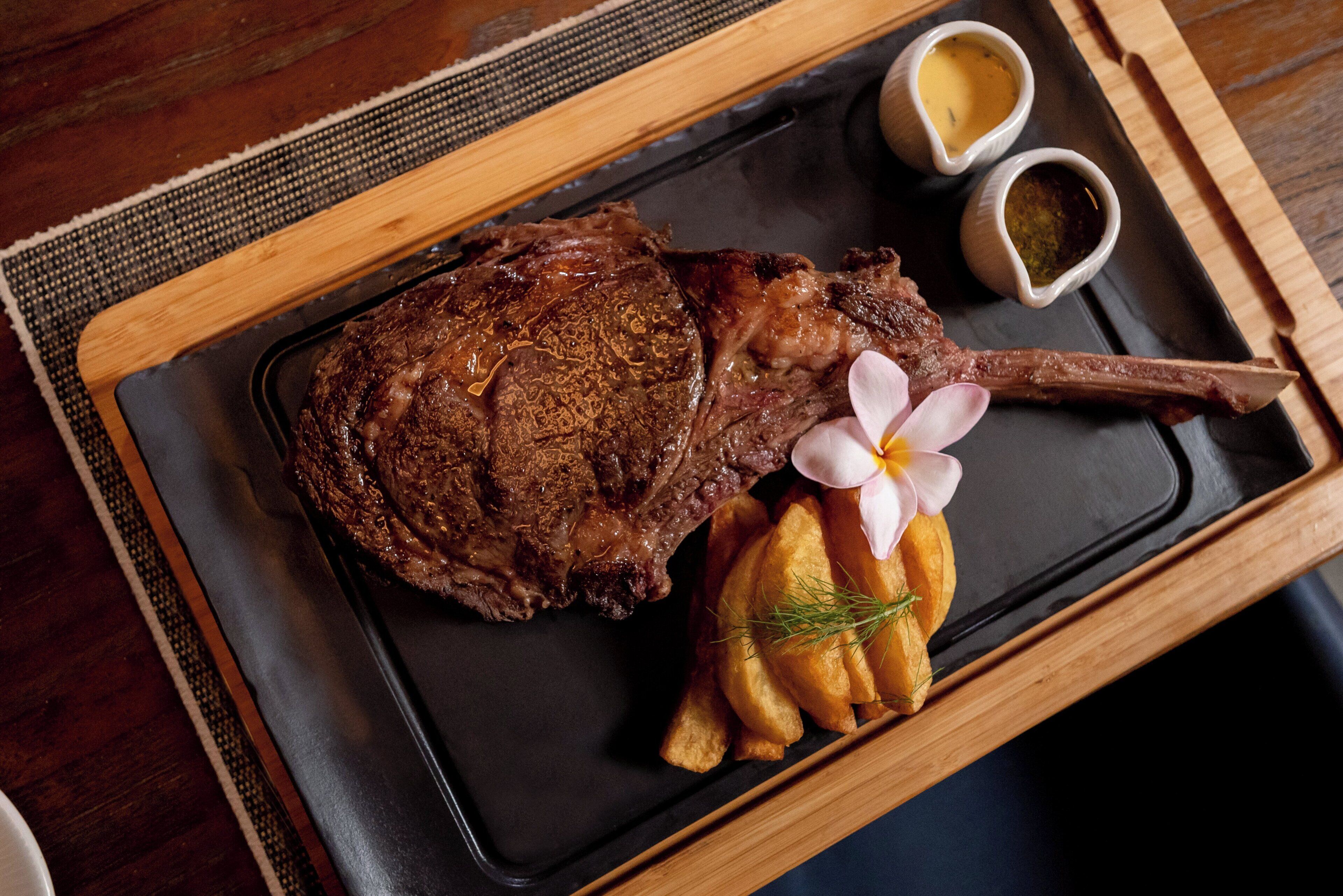 A steak with a flower garnish on a black plate, accompanied by two small sauce containers and a side of fries.