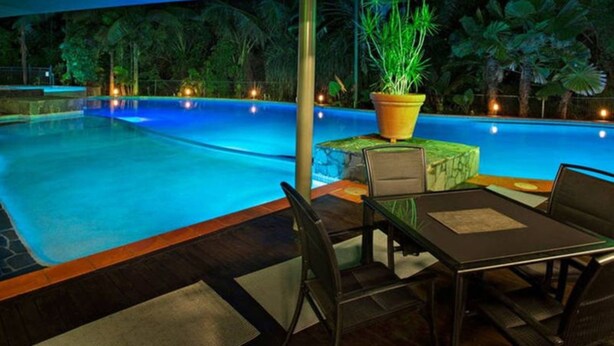 Zona de piscina exterior con mesa y sillas, rodeada de abundante follaje y un toldo azul.