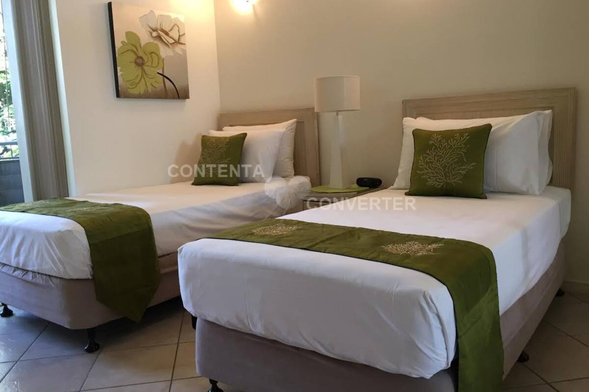 A hotel room with two beds, each with white bedding and green throw pillows. There is a painting on the wall and a lamp on the nightstand.