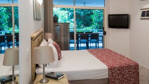 Habitación de hotel con una cama, una mesita de noche con lámpara, un televisor montado en la pared y vistas al exterior a través de amplios ventanales.