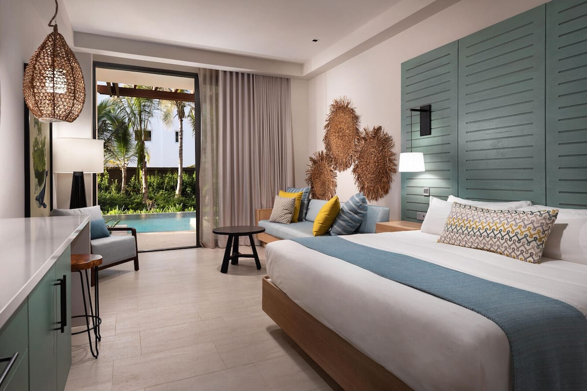 A modern hotel room with a bed, a sofa, a small table, and a view of an outdoor pool.