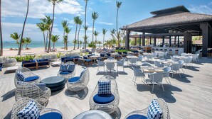 A beachside resort with outdoor seating areas, palm trees, and a thatched-roof structure.