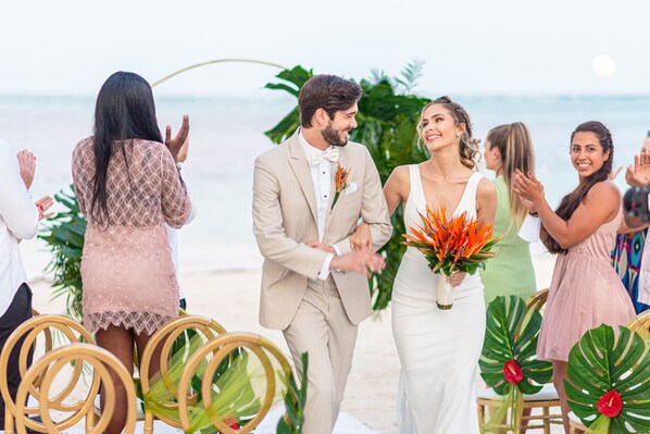 A beach wedding ceremony with guests seated on wicker chairs, a man in a beige suit, and a woman in a white dress holding a bouquet.