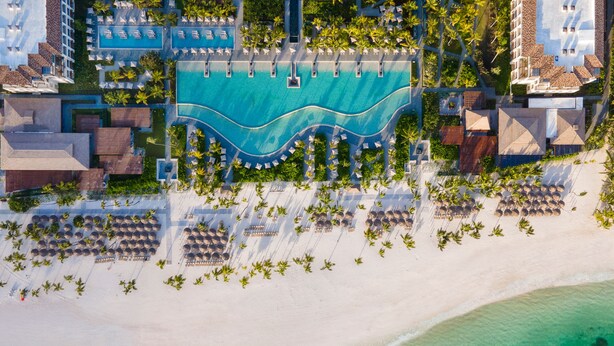 An aerial view of a resort with a large swimming pool, beachfront seating, and palm trees.