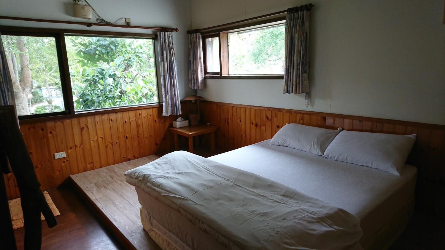 A room with a bed, wooden walls, a small table, and windows with curtains.