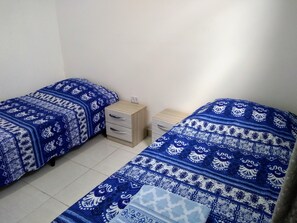 Quarto com duas camas, cada uma com colcha estampada azul, um pequeno criado-mudo de madeira com gavetas e uma parede branca.