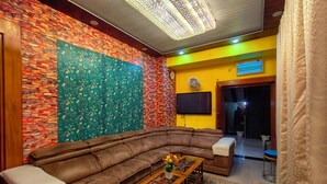 Een woonkamer met een bruine leren bank, een houten salontafel met een bloemist centraal stuk, een kamerkleed met dessin, een bakstenen muur met wandkleed met bloemen, en een gele wand met een klok en een televisie.
