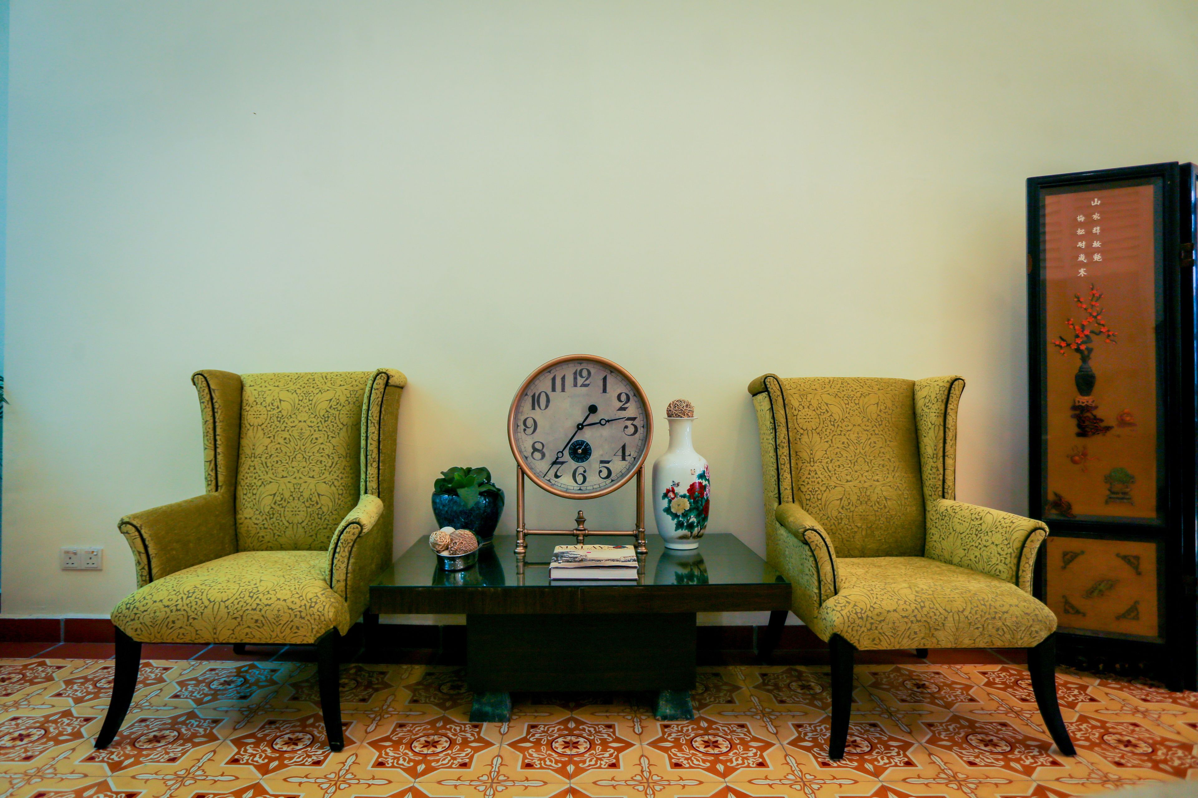 A room with two armchairs, a round table with a clock, a vase, and a framed picture on the wall.