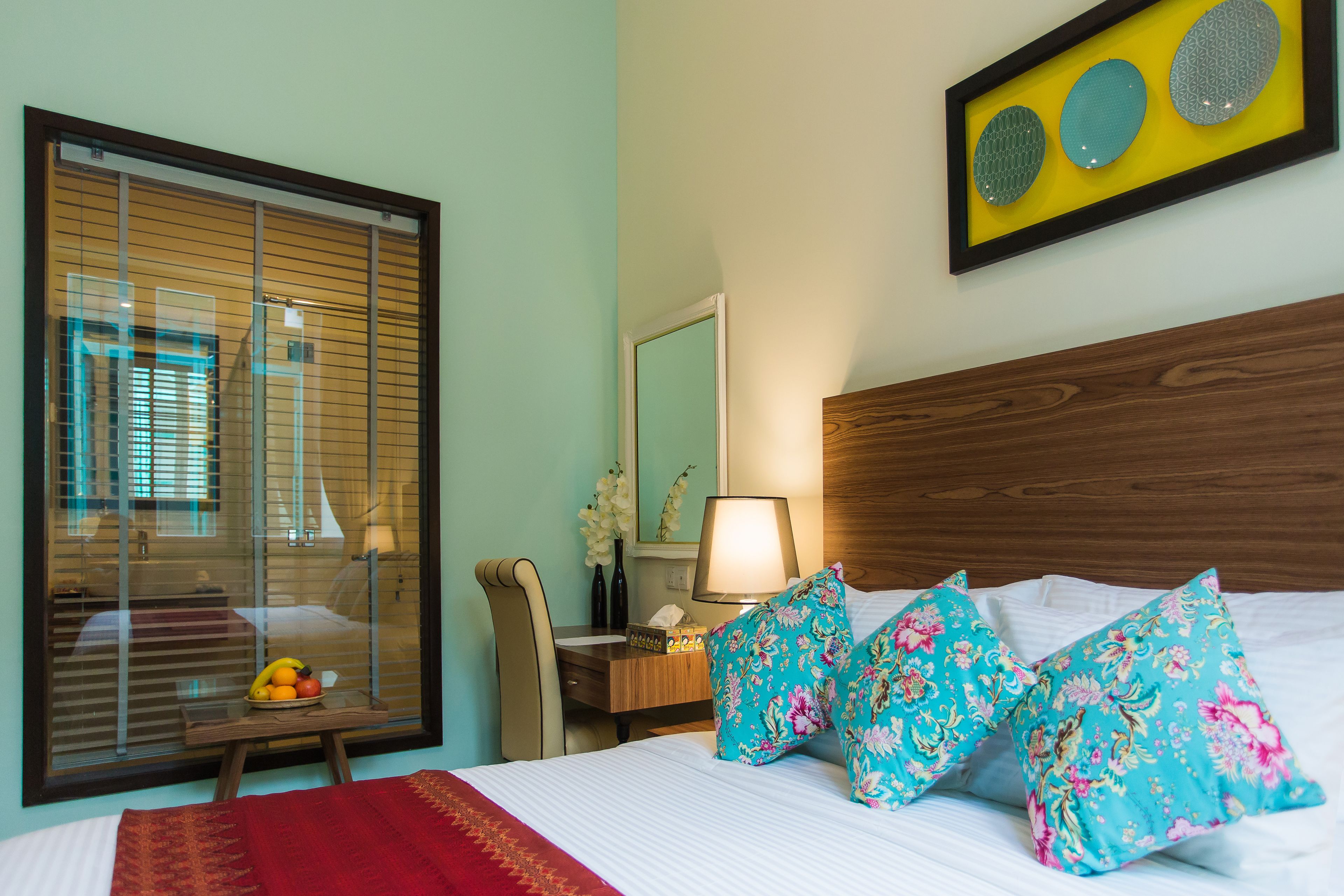 A hotel room with a large window, a wooden headboard, a bedside table with a lamp, and floral pillows.
