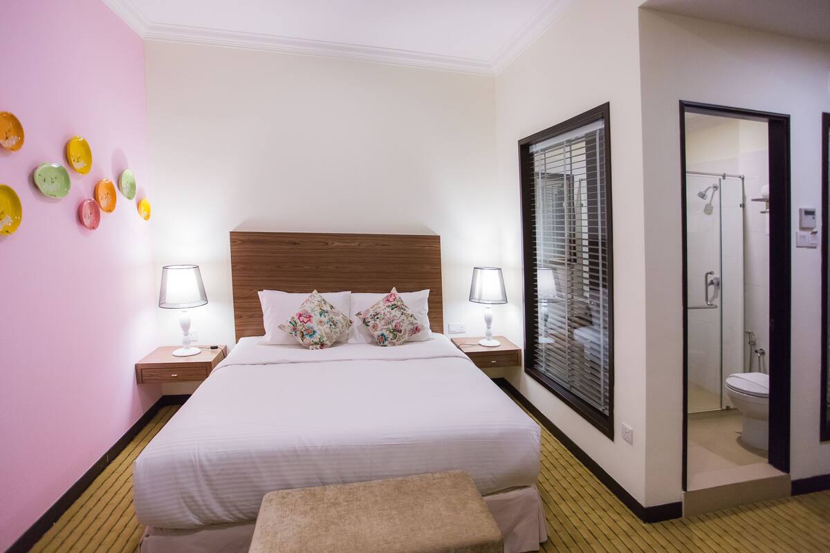 A hotel room with a large bed, two bedside tables with lamps, a wooden headboard, a small bench, a bathroom with a shower, and a glass door leading to the bathroom.