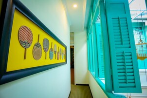 Un couloir orné d’une collection de raquettes de badminton encadrées sur un mur jaune, une fenêtre turquoise avec des volets et un plancher en bois.