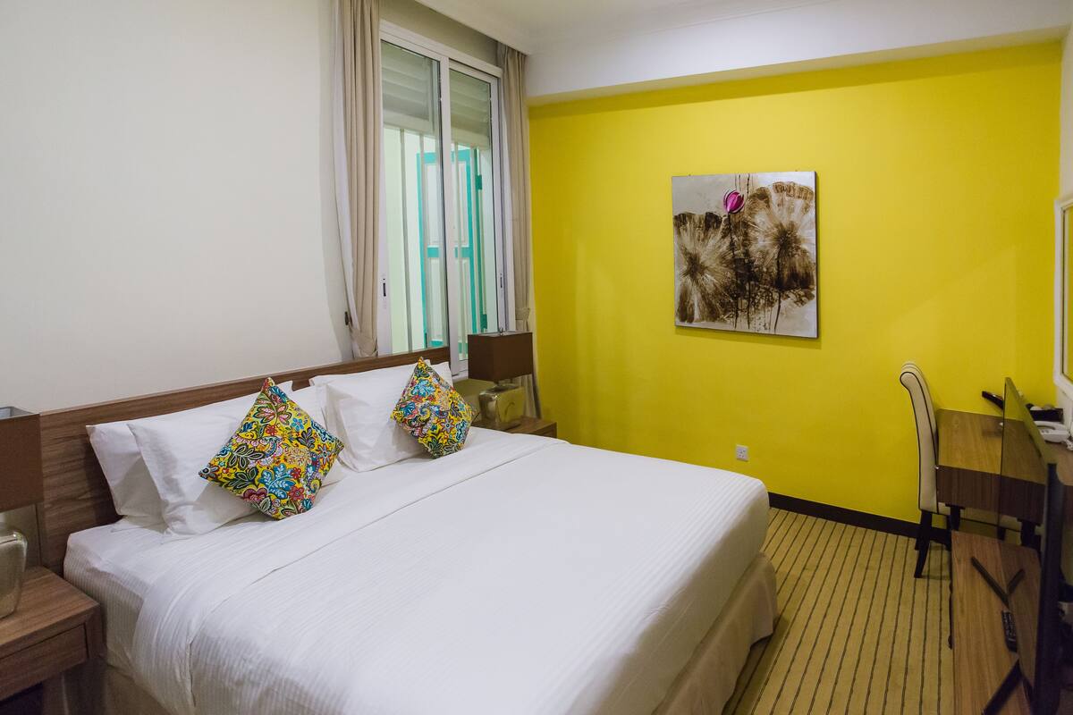 A hotel room with a bed, a desk, a chair, a window with curtains, and a painting on the wall.