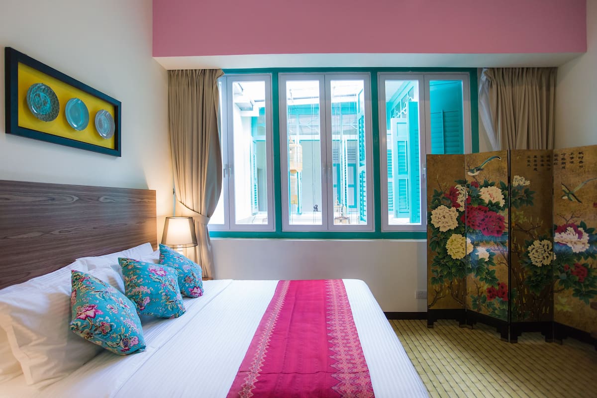 A neatly made bed with floral pillows, a pink bedspread, and a view of a building through a window with blue shutters.