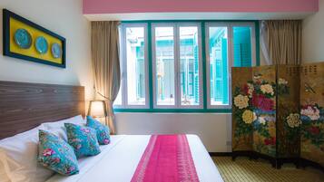 A neatly made bed with floral pillows, a pink bedspread, and a view of a building through a window with blue shutters.